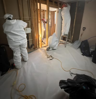 A crew in PPE stripping the room to the studs inside the poly-lined containment, with bagged waste staged