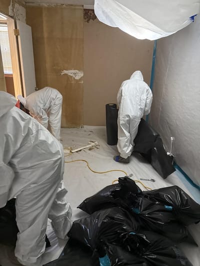 Workers in protective suits bagging waste inside a plastic-lined asbestos containment area
