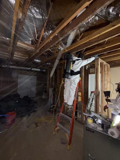 A technician with a backpack HEPA vacuum cleaning the exposed ceiling framing of a stripped basement during abatement