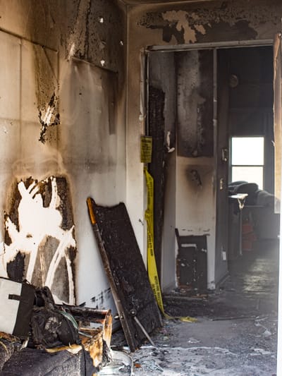A burned-out hallway with soot-blackened walls, charred debris on the floor, and caution tape in the doorway