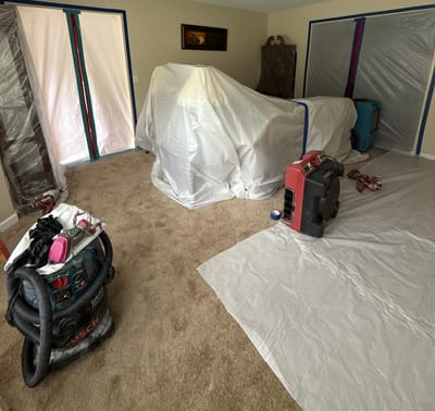 A carpeted room sealed with plastic containment sheeting, furniture covered and a shop vacuum and air mover set up for mold removal