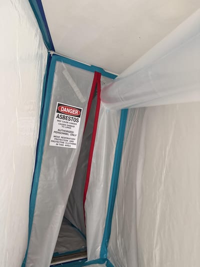 Sealed asbestos containment with a zippered entry, warning sign, and negative-air duct running in