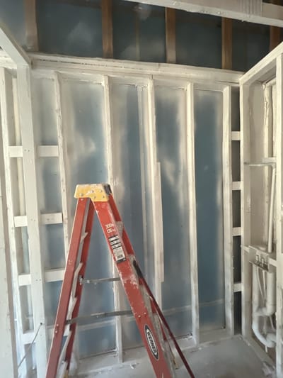 Exposed wall framing sealed with a white encapsulant coating, with a step ladder in the room