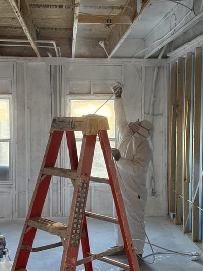 A technician in protective gear spraying sealant onto the ceiling framing of a fire-damaged room stripped to the studs