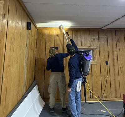 Two technicians HEPA-vacuuming the ceiling and wood-paneled walls of a room, one wearing a backpack vacuum
