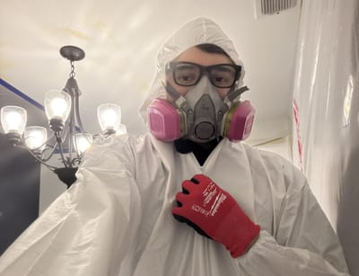 A restoration technician in a white hazmat suit, half-face respirator, goggles and red gloves inside a plastic-sheeted containment area