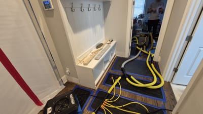 Injection drying mats and hoses taped across a hardwood floor, with plastic containment sheeting on the wall