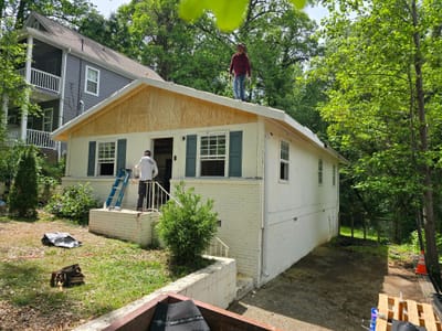 Two workers rebuilding a house roof, one on the fresh decking and one on a ladder, with new plywood sheathing on the gable
