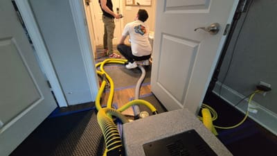 Two technicians drying a hardwood floor with yellow injection-drying hoses taped across the boards, running to drying equipment