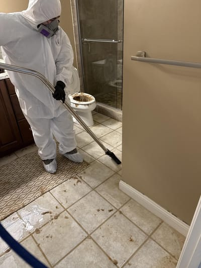 A technician in a protective suit and respirator cleaning a tiled bathroom floor after a sewage toilet overflow