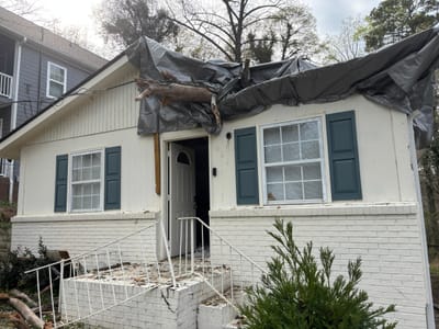 A fallen tree limb punched through the roof eave of a house, with a tarp draped over the damaged section