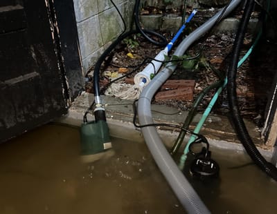 A submersible sump pump sitting in murky floodwater in a basement corner, with discharge hoses running along a concrete block wall