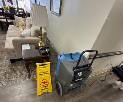 A resident common room with a sofa and lamp, a dehumidifier running on the floor, and a bilingual Caution Wet Floor sign