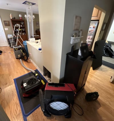 A unit being dried: air movers and a dehumidifier on the hardwood floor, a drying mat, and a clear duct carrying airflow up to a ceiling vent