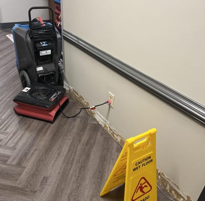 A hallway during drying: a dehumidifier and a low air mover against the wall with the baseboard removed, a Caution Wet Floor sign in front, and the walkway kept clear