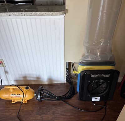An injection drying setup: a yellow drying unit with a manifold of hoses feeding air into a wall cavity, next to a ducted air mover, drying the wall without opening it up
