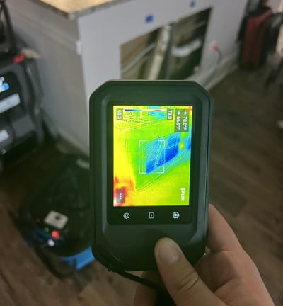 A handheld thermal imaging camera held up to a wall, its screen showing cool blue areas of moisture against warmer tones, in a unit being dried
