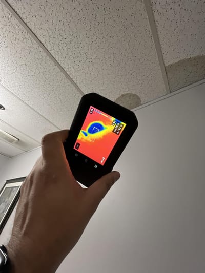 A thermal imaging camera pointed up at a stained drop-ceiling tile, its screen showing a cool blue cold spot where moisture has collected above the ceiling